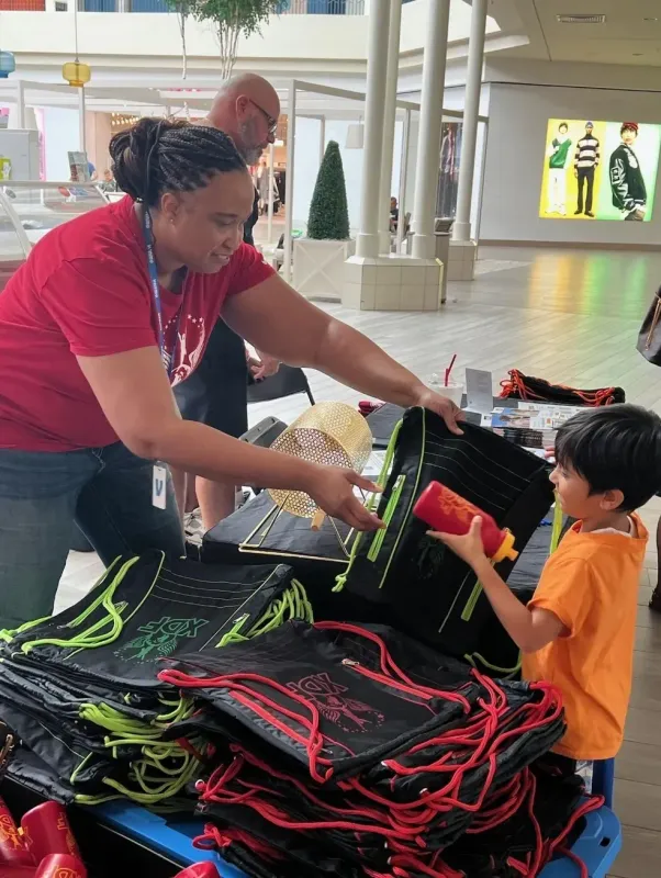 An instructor in a red shirt helping a young child in an orange shirt examine items at an event table display indoors.