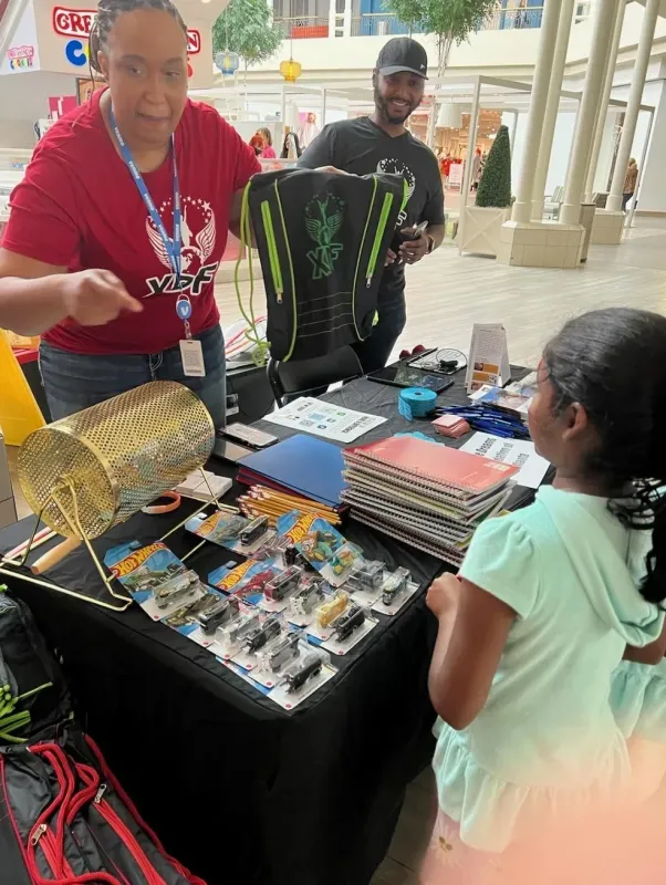 Community education event at an indoor mall with organizers in red and black shirts demonstrating gear and materials to attendees at a black table display.